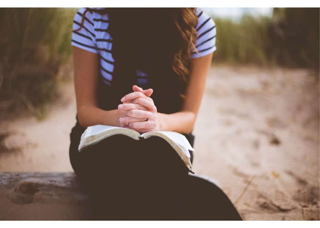 Woman sitting on a log outside praying over open Bible.