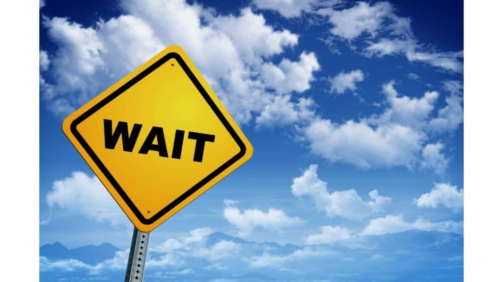 Image of yellow diamond-shaped road sign that says "WAIT" against a blue sky with white clouds.