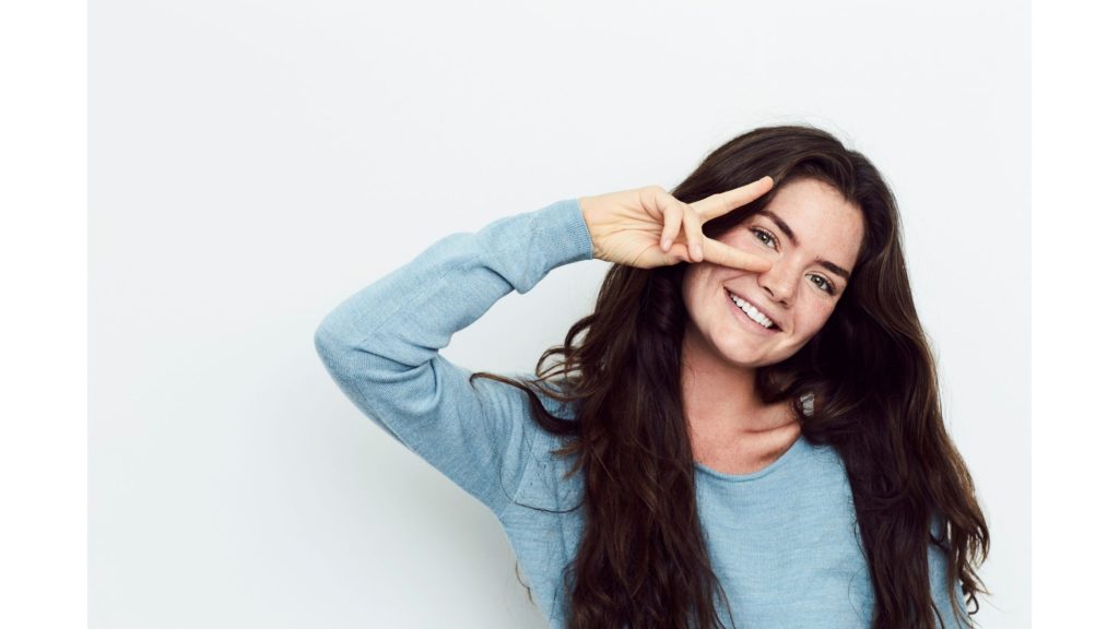 Woman with long dark wavy hair in a blue sweater flashing a peace sign.