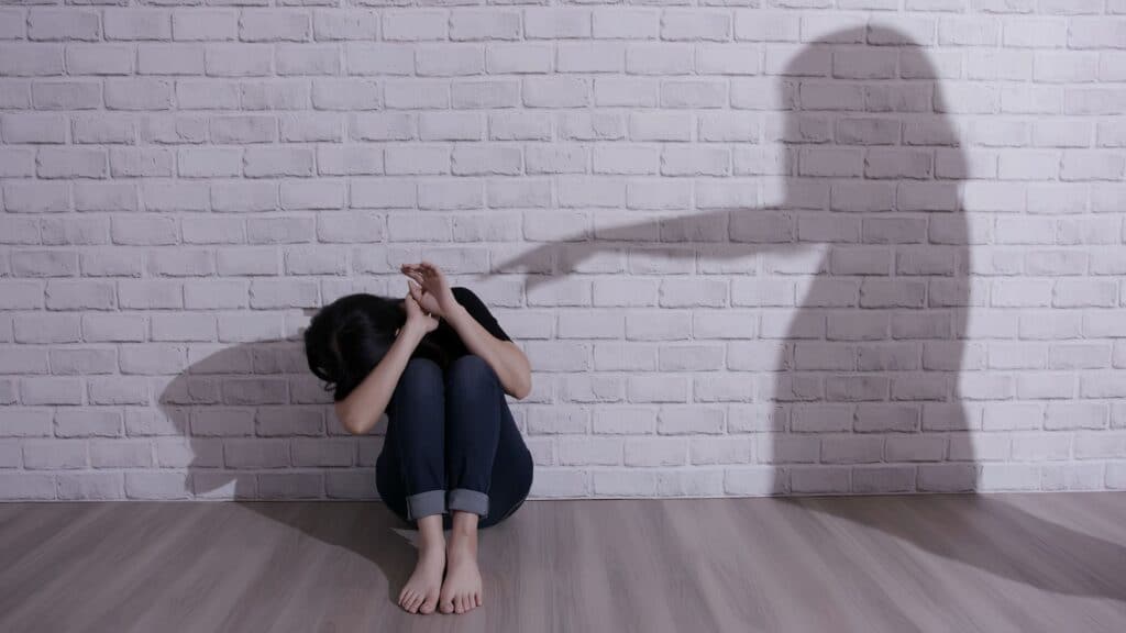 Shadow on a white brick wall pointing at, or accusing, a woman in a defenseless position on the floor.
